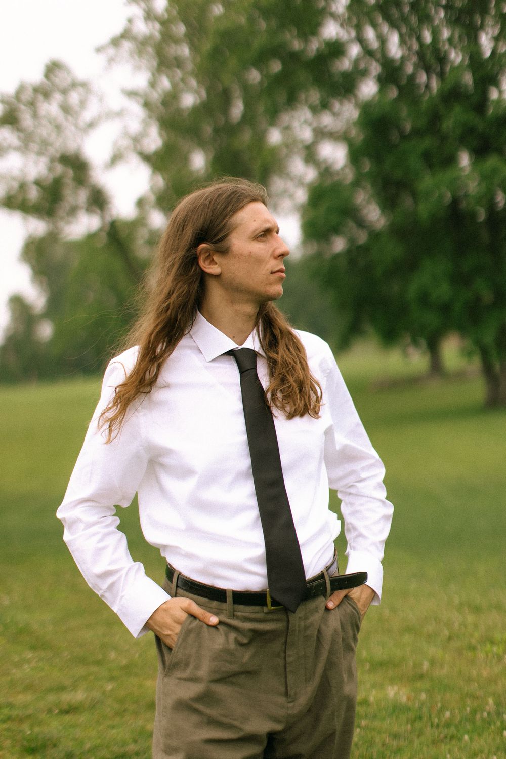 Person in formal attire with long hair and tie poses in grassy outdoor setting.