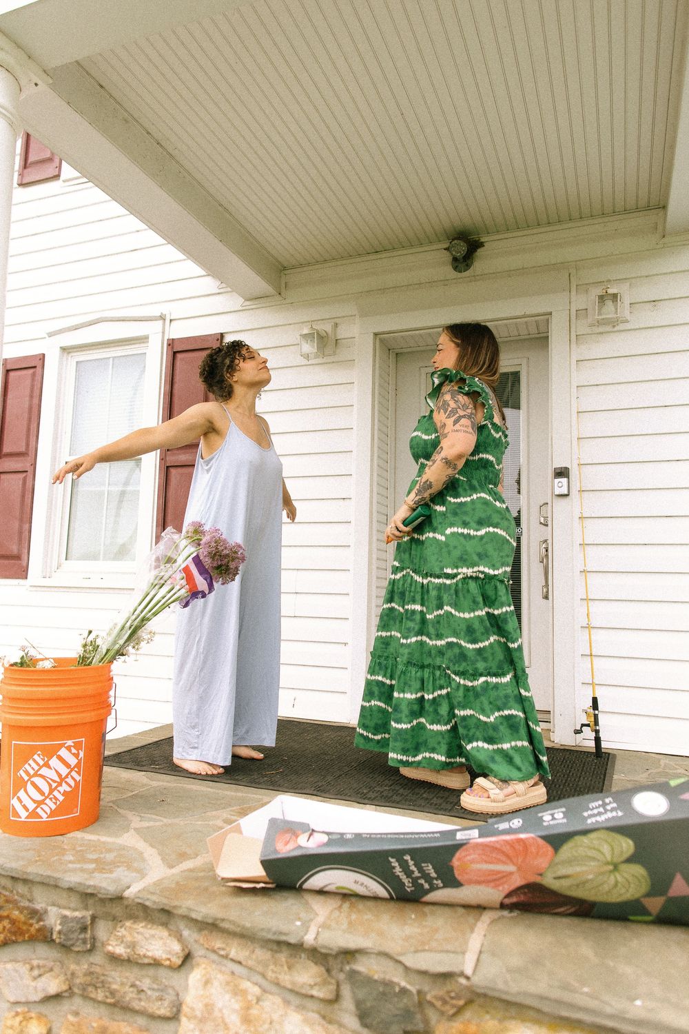 Two people in contrasting dresses stand on a porch with a Home Depot bucket and decorative rug.