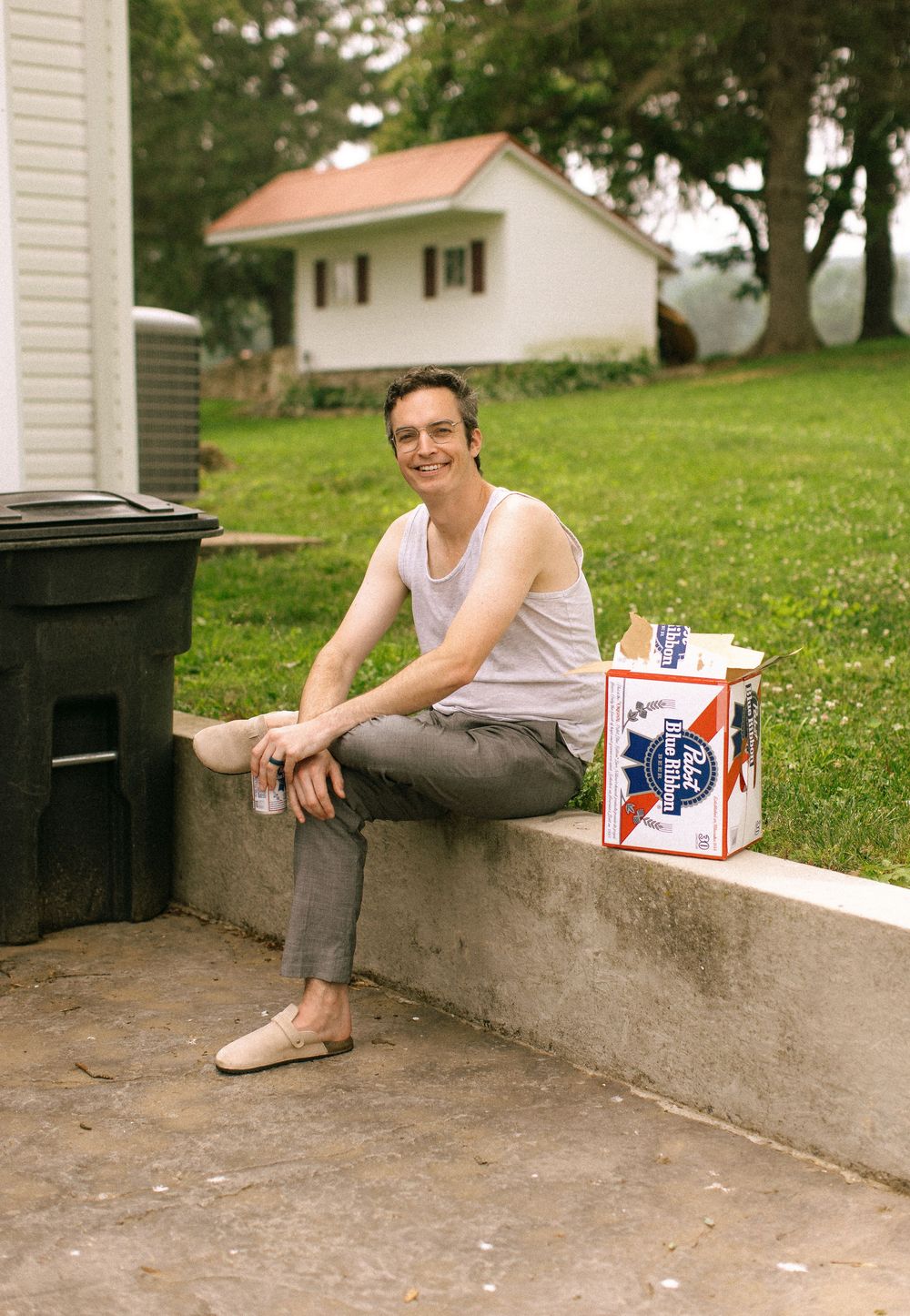 A person sits casually on a concrete ledge outdoors with a Pabst Blue Ribbon beer can nearby on a sunny day.