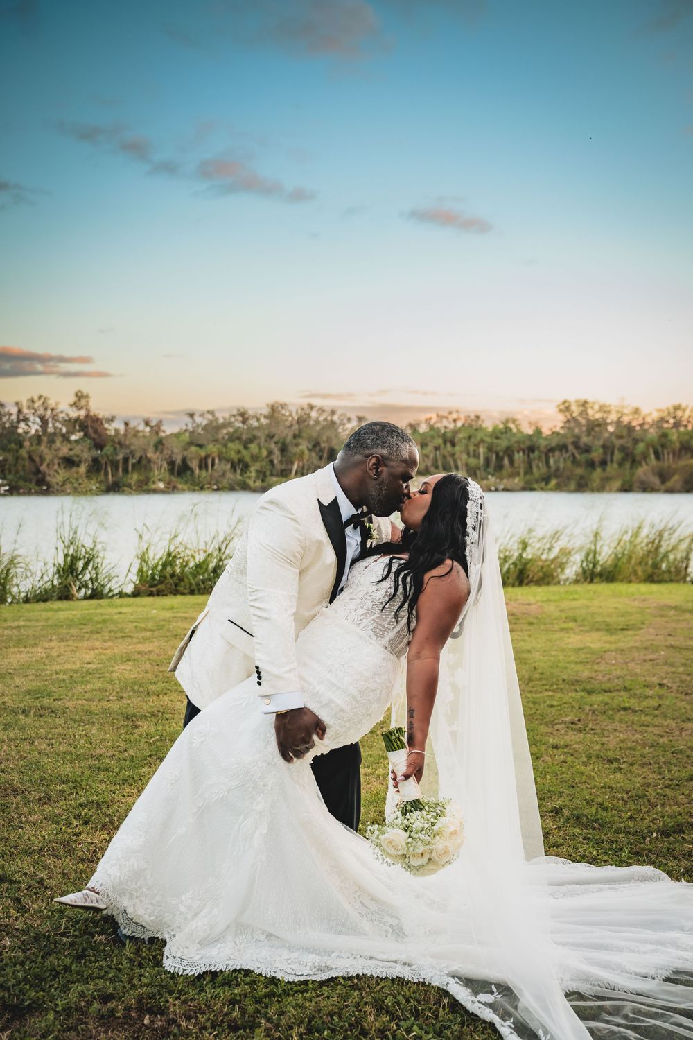 A couple shares a romantic kiss in wedding attire by a lakeside at sunset, bride holding white bouquet with long veil. wedding photography