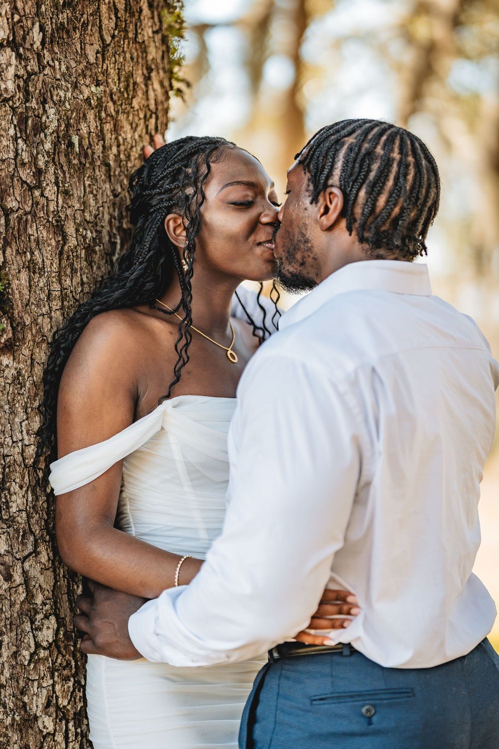 A Black couple shares a romantic kiss in wedding attire near a tree, bride in white off-shoulder gown. Tampa wedding photography