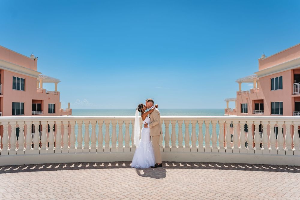 Bride and groom kissing on a pink resort balcony overlooking the turquoise ocean under a clear blue sky. Hyatt Regency Clearwater, Photo