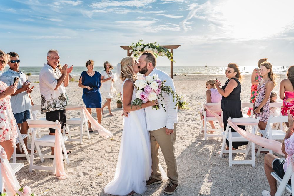 Bride and groom share first kiss on sandy beach wedding ceremony with guests cheering and floral arch backdrop. Wedding Photography, Photo