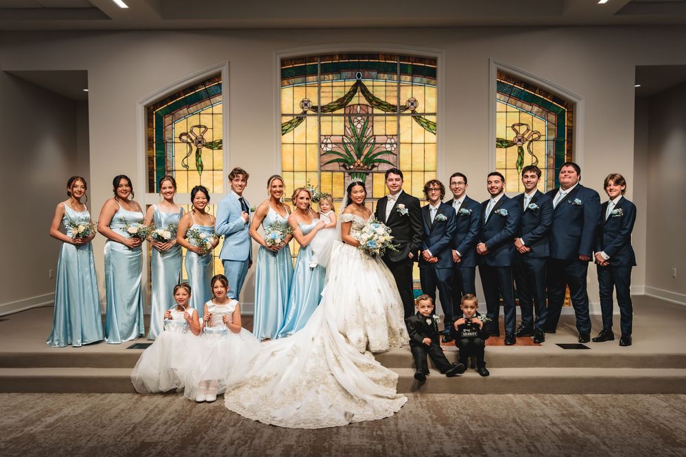 Wedding party poses in chapel with stained glass windows, bride in lace gown, bridesmaids in blue dresses, groomsmen in navy suits. Photo