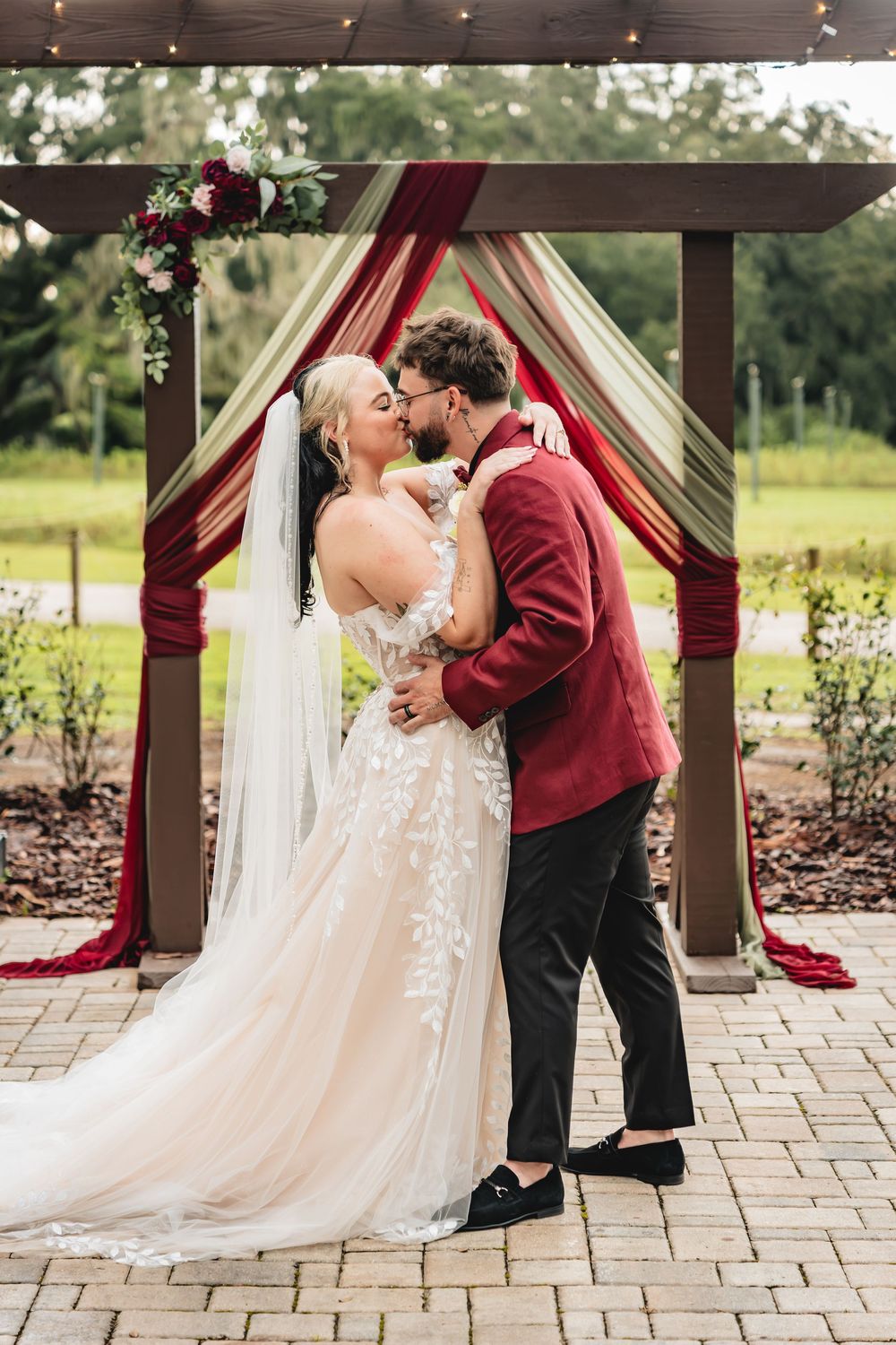 Bride and groom share first kiss under decorated wooden arch with burgundy draping at outdoor wedding ceremony.