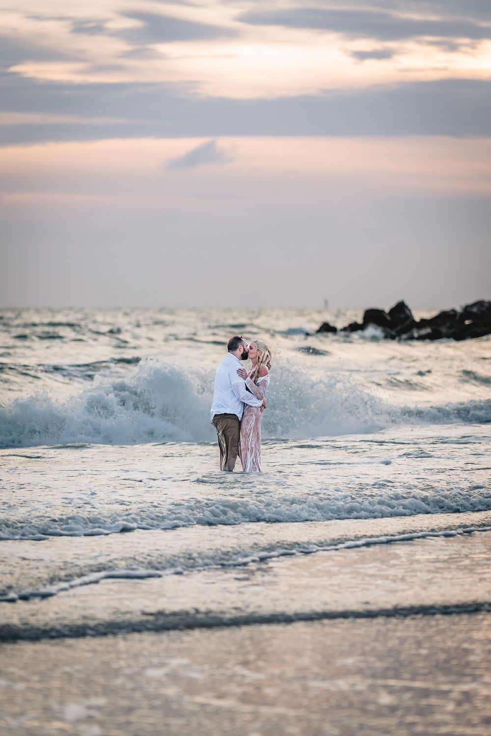Couple embracing in ocean waves at sunset on a beach, wearing romantic attire with colorful sky backdrop. Wedding Photography, Photographer