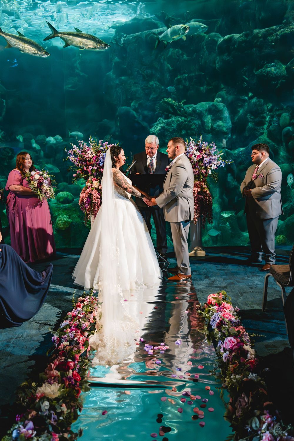 Couple exchanges vows at an aquarium wedding ceremony surrounded by colorful floral arrangements with sharks swimming overhead. Tampa Photo