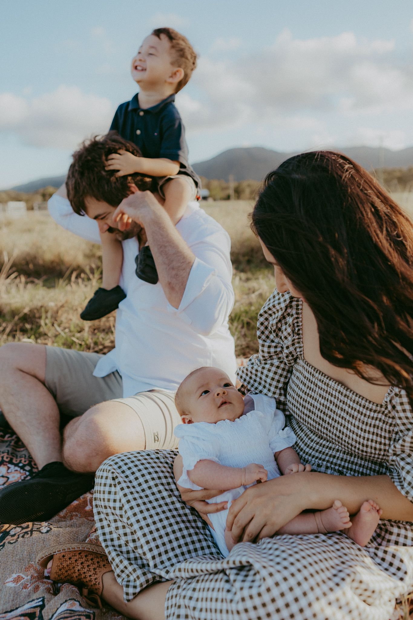 Family of four enjoys playful outdoor picnic in field, father lifting toddler while mother holds baby.