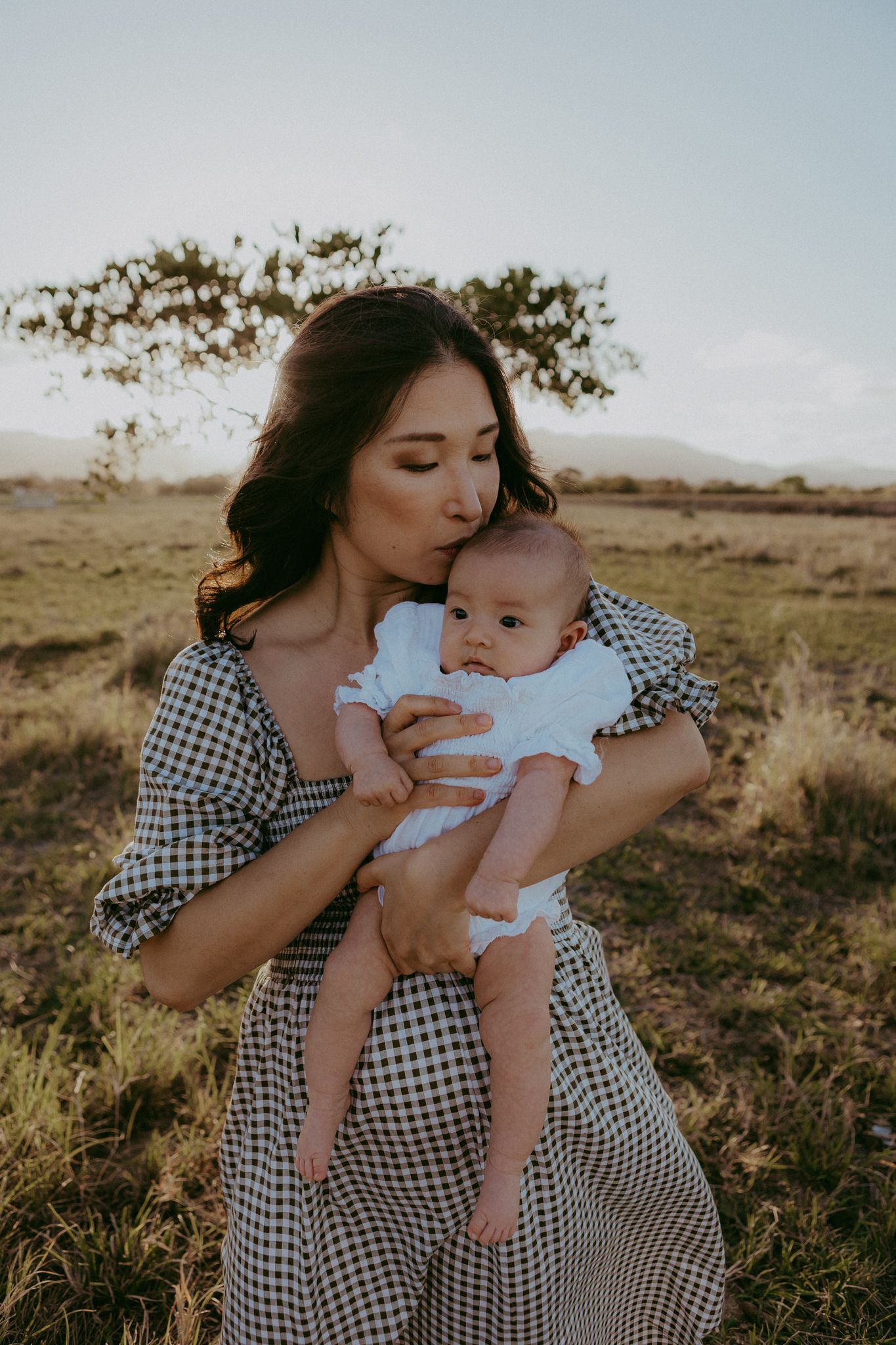 Mother in gingham dress kisses newborn baby while standing in open grassy field at sunset.
