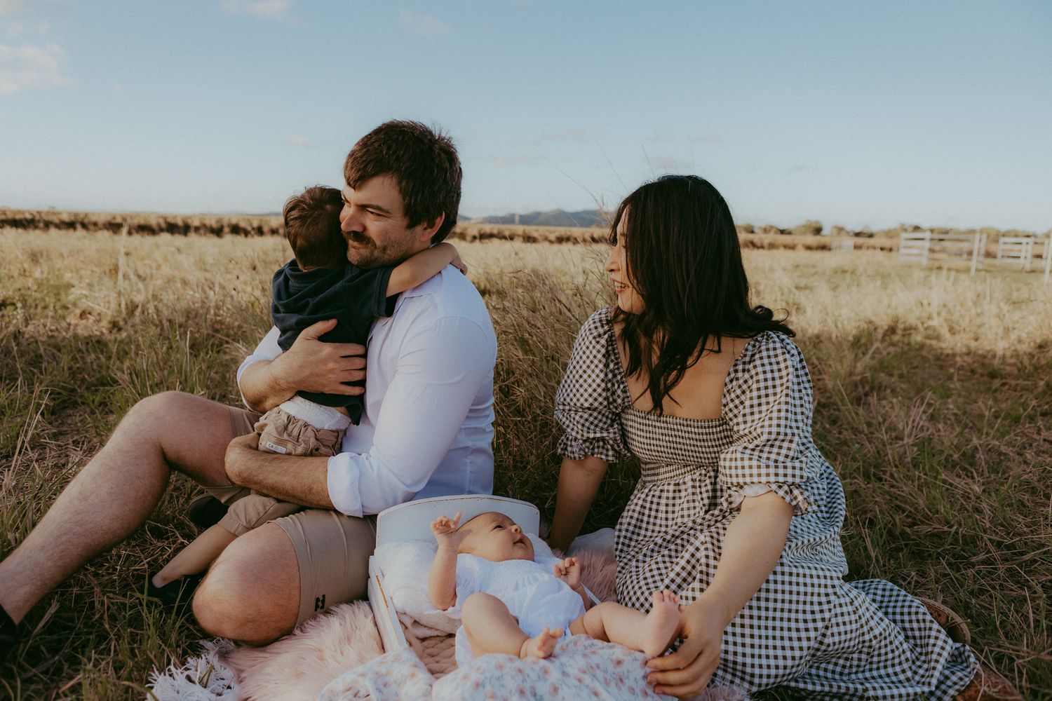Family sitting on blanket in golden field, father holding toddler while mother in gingham dress smiles.