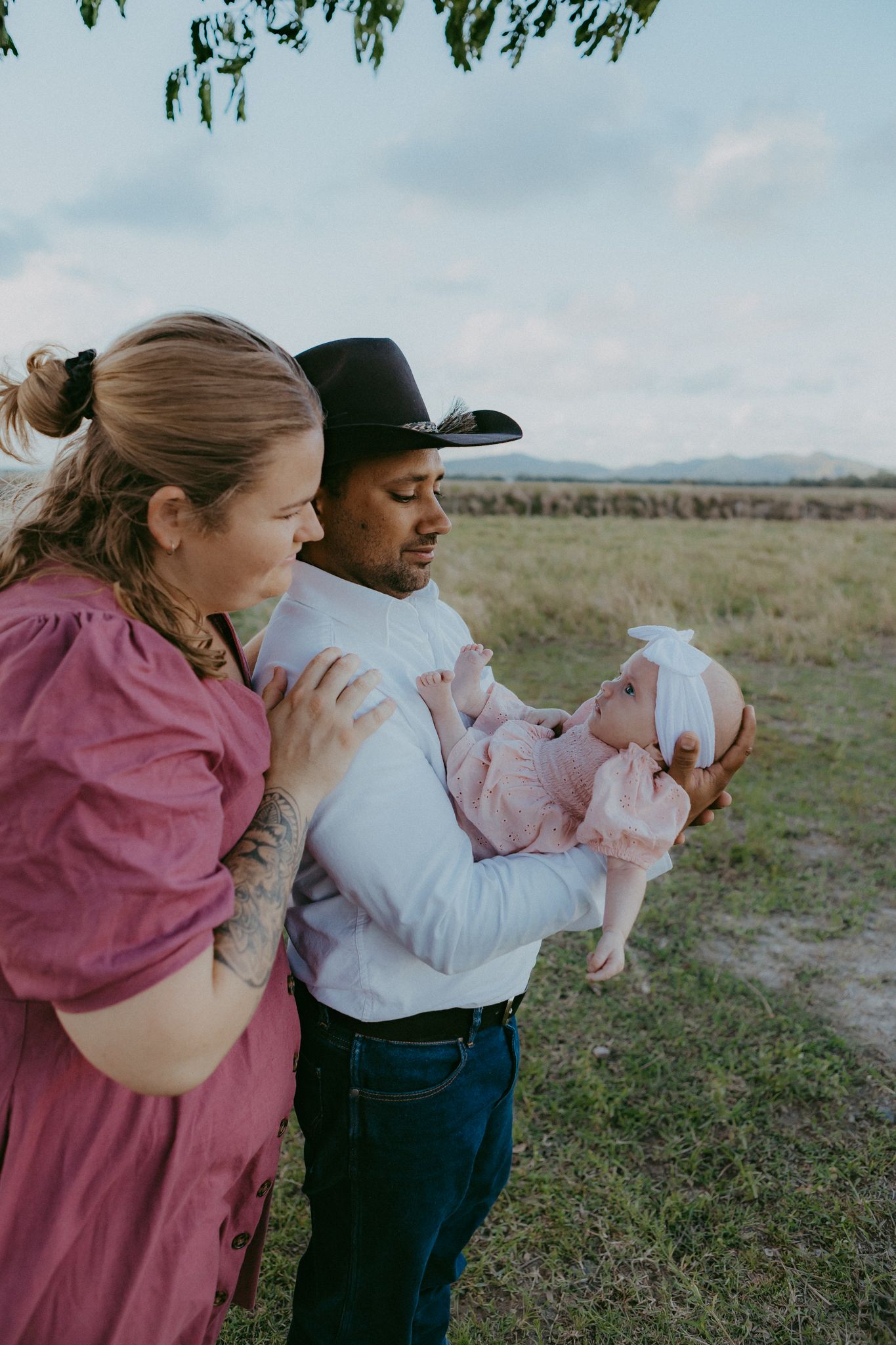 Parents in field admiring newborn baby in pink dress held by father wearing black cowboy hat.