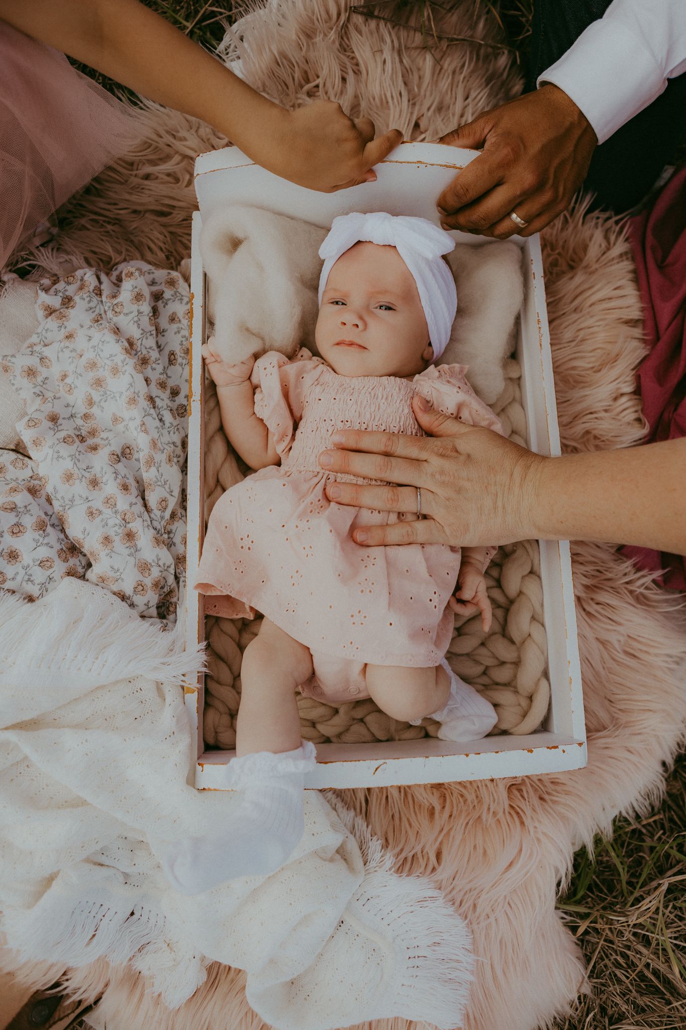 Newborn baby in pink dress and white headband lying in white wooden box on fluffy blanket.
