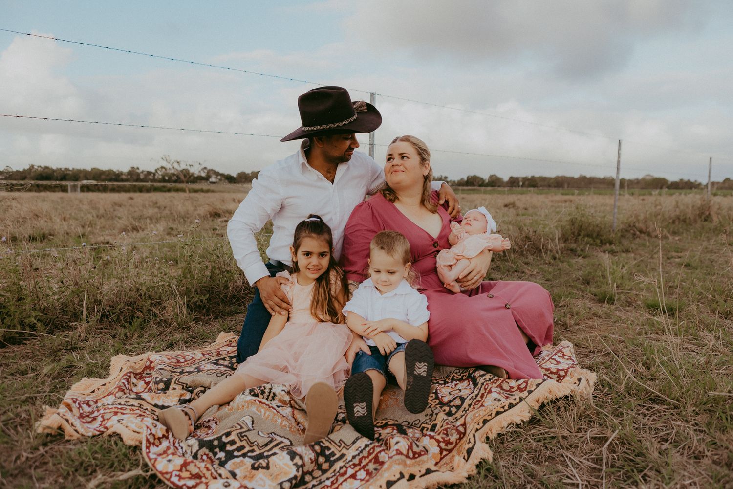Family of four sitting on patterned blanket in field, father in cowboy hat holding newborn baby.