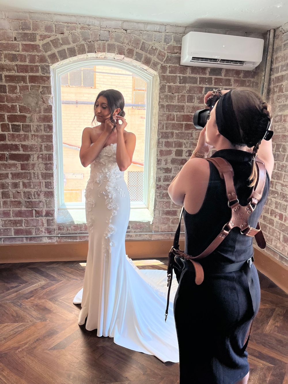 Behind-the-scenes sequence showing a photographer in black capturing a white dress display against exposed brick walls.