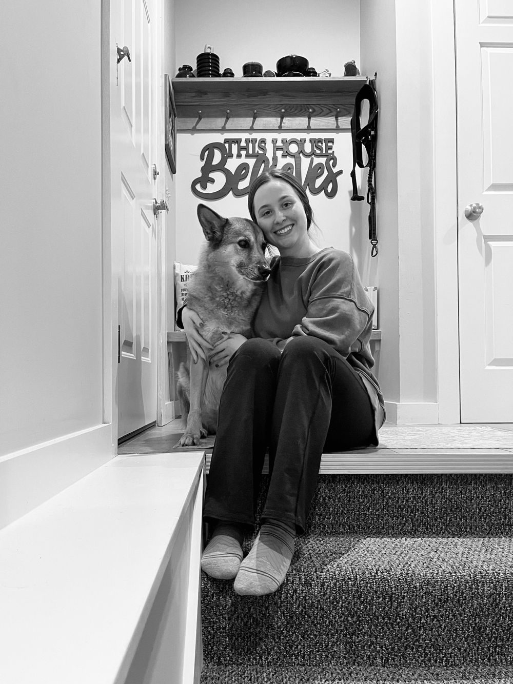 Black and white portrait of a person sitting with their German Shepherd dog on carpeted stairs.