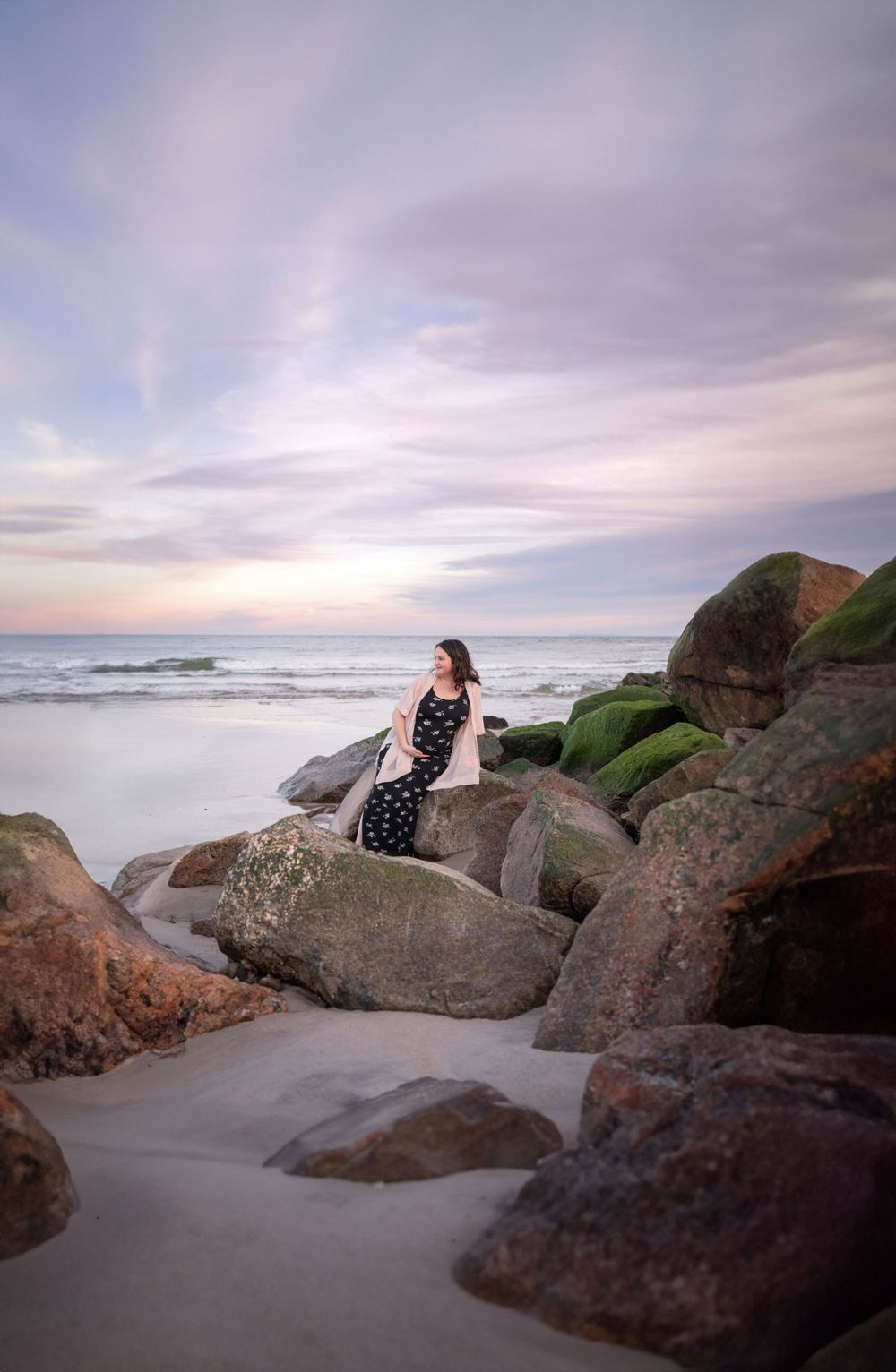 Couple sitting on coastal rocks at sunset with ocean waves in background.