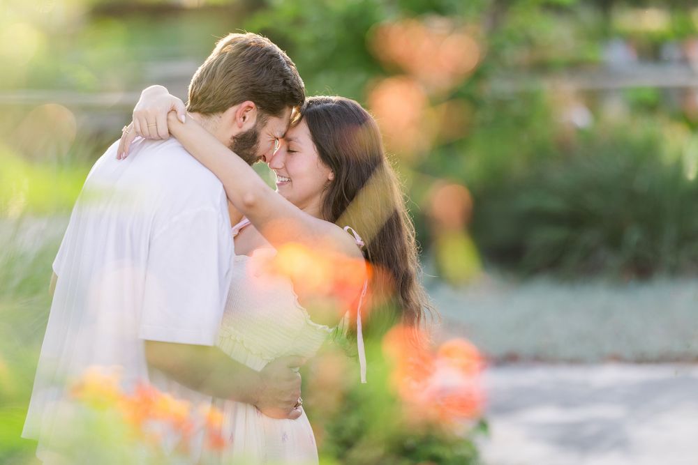A romantic couple shares tender kisses in a garden setting with soft natural lighting and blurred floral elements.