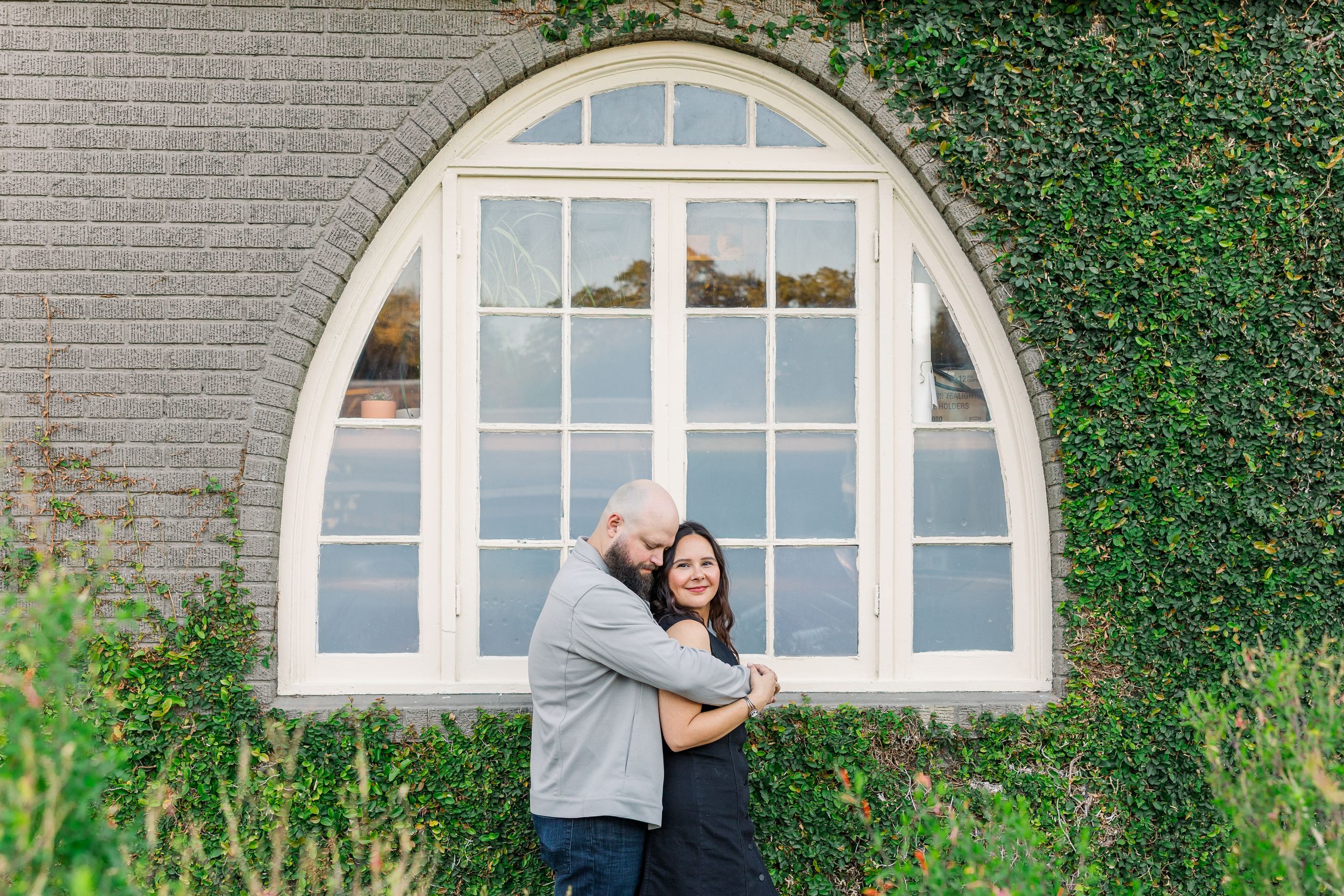 A couple embraces in front of a brick wall with arched windows and climbing ivy.