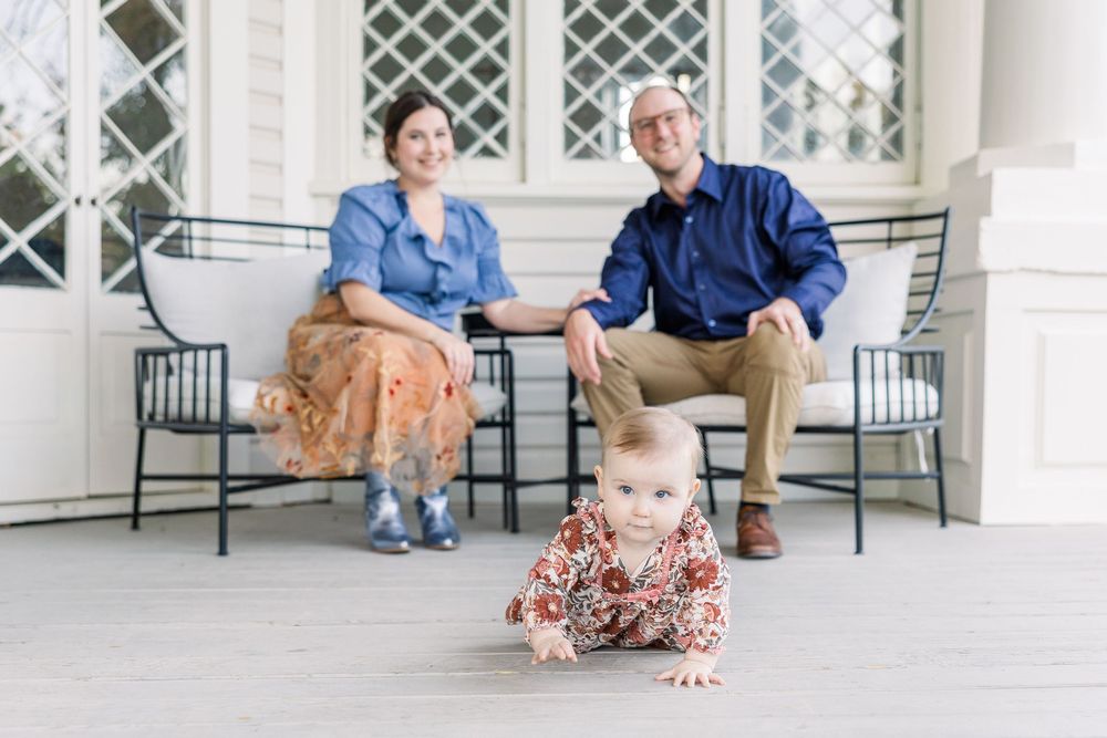 A baby in floral dress crawls on a porch while two adults sit on metal chairs behind.