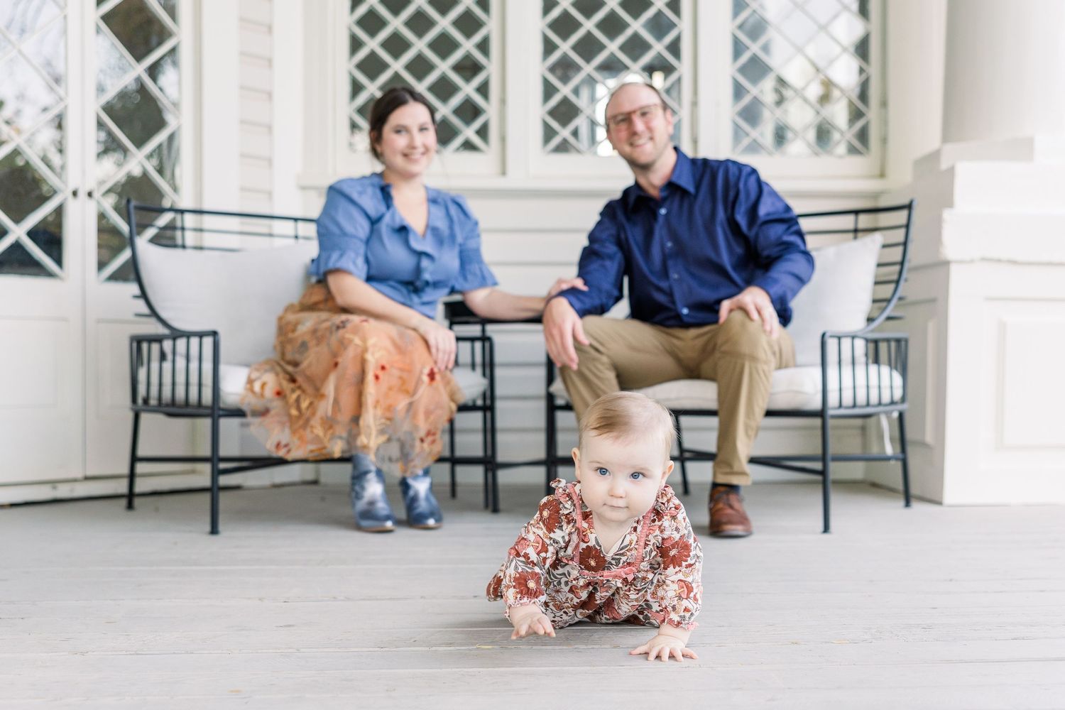 A baby in floral dress crawls on a porch while two adults sit on metal chairs behind.