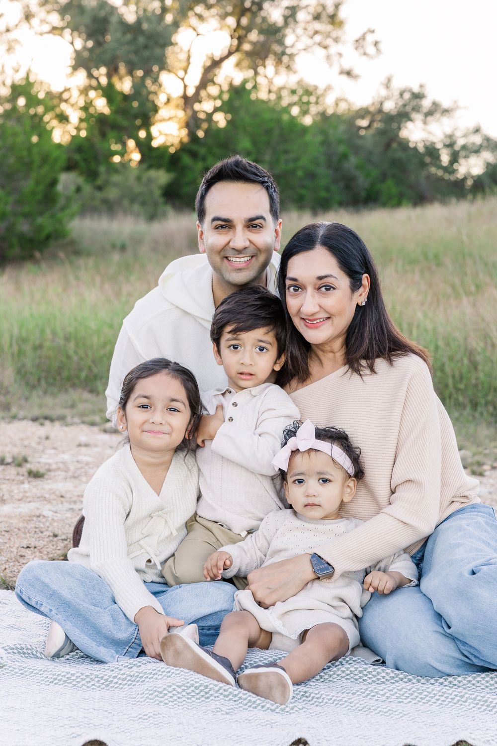 Family relaxes together on a white blanket outdoors during a casual photo session in a natural field setting.