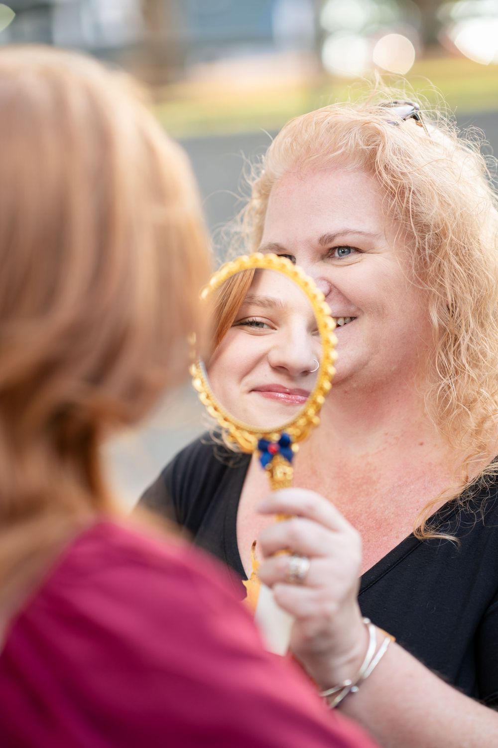 Someone looking at their reflection through an ornate handheld gold mirror while smiling outdoors with bokeh background.