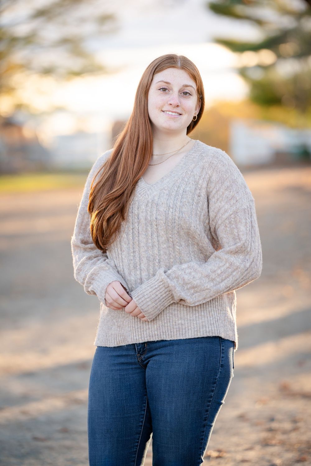 A person wearing a cozy beige sweater and blue jeans poses outdoors during golden hour lighting.