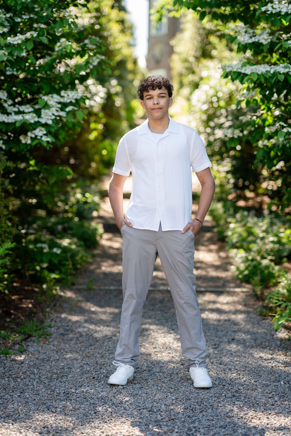 A person wearing a white button-down shirt and gray pants stands on a gravel path surrounded by lush green foliage.