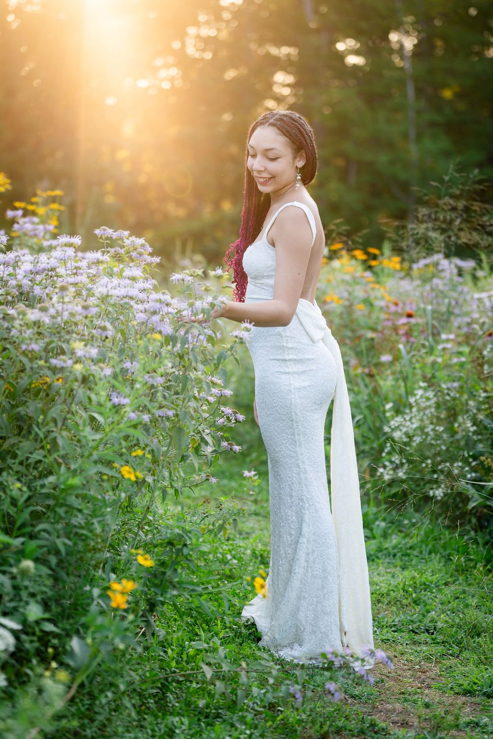 Young woman in white lace gown standing among wildflowers in golden sunset light in a lush garden.
