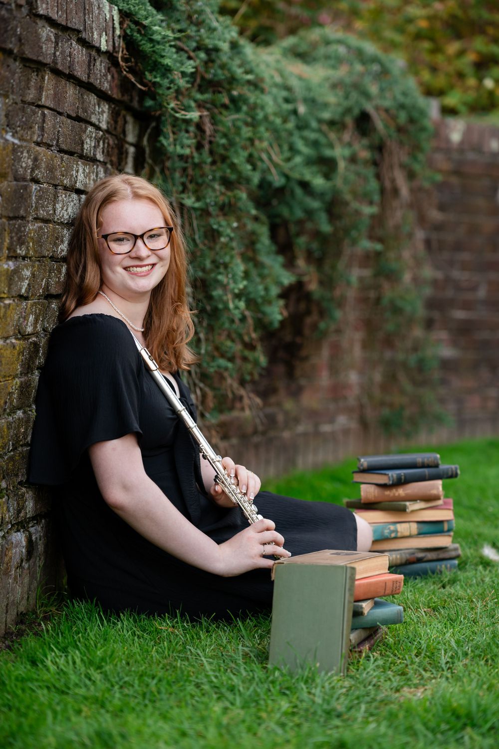 A person in a black dress sits against a brick wall with a stack of books on grass in an outdoor setting.