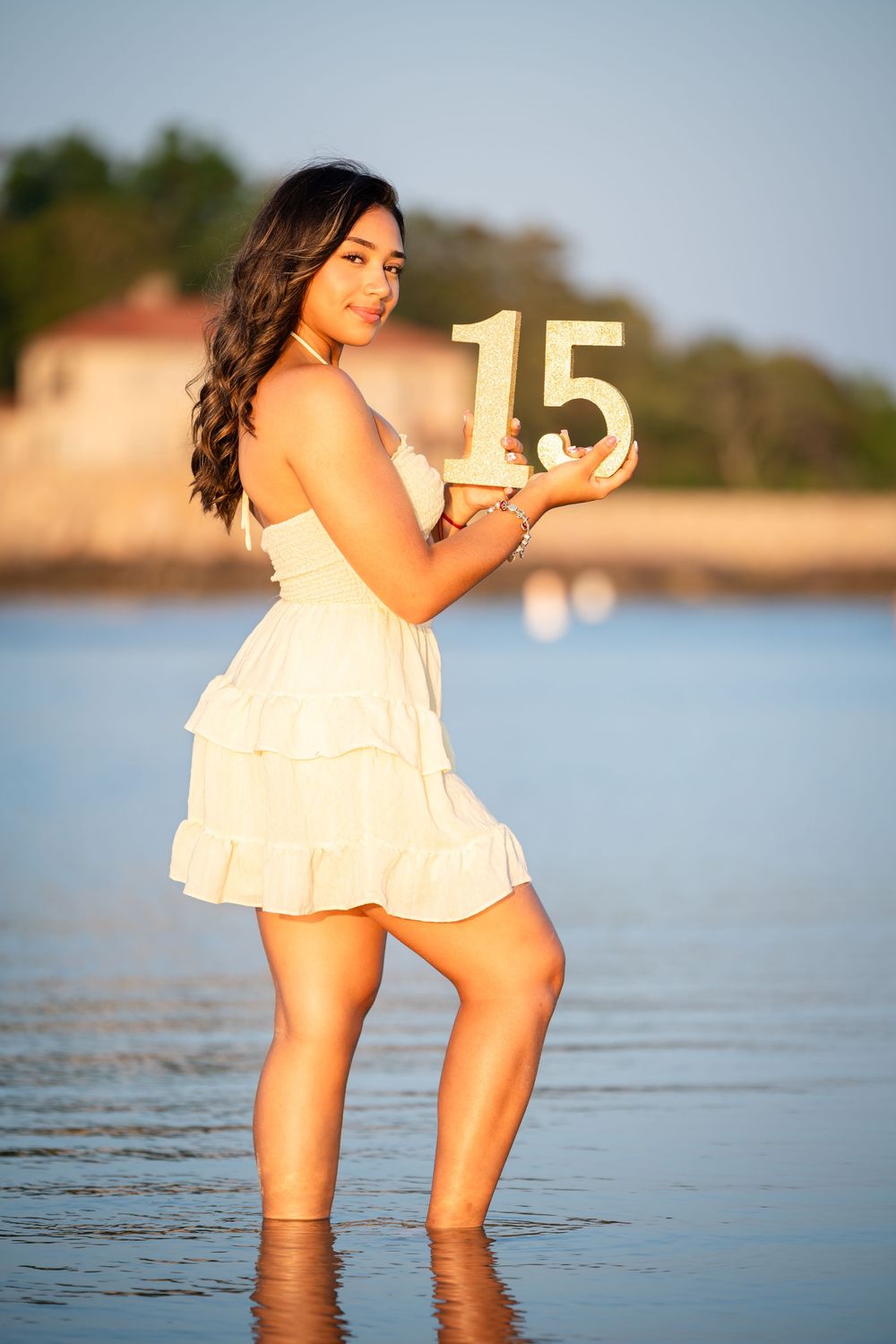 A white dress-wearing person stands in shallow water holding gold number 15 during sunset at the beach.