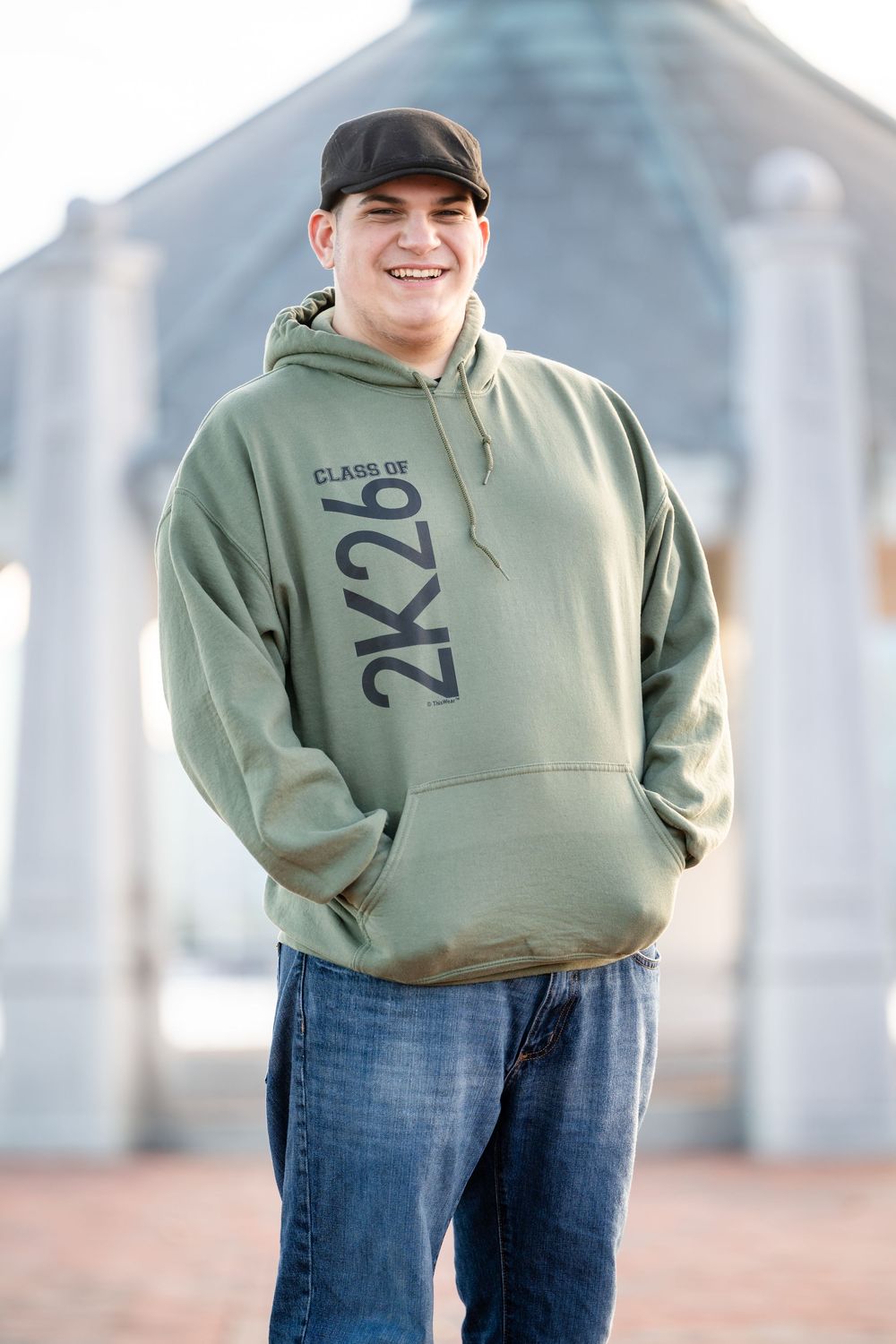A person wearing a green 2k2b hoodie and baseball cap stands smiling outdoors with a building in the background.
