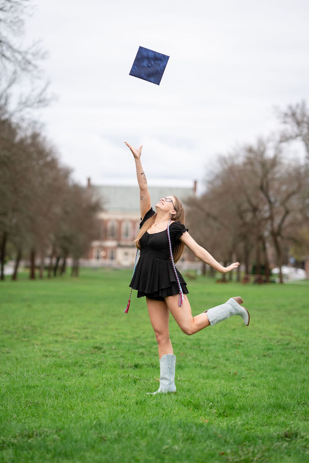 Graduate in black dress and blue socks tosses cap in celebration on green lawn with historic building in background.