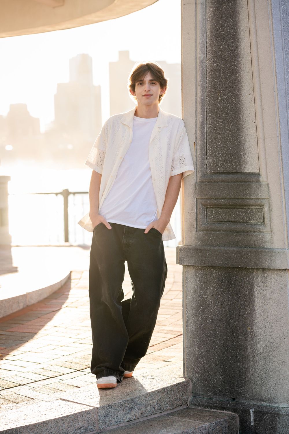 A person in white t-shirt and black pants leans against a stone column at sunset with city buildings in background.