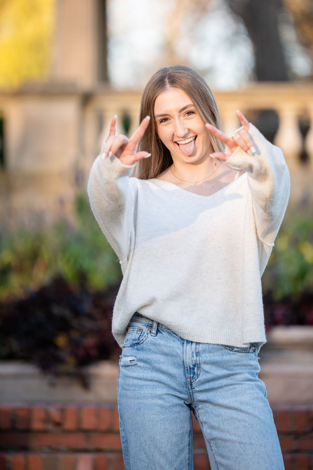 Person in light colored sweater and blue jeans poses playfully against a brick wall during golden hour.