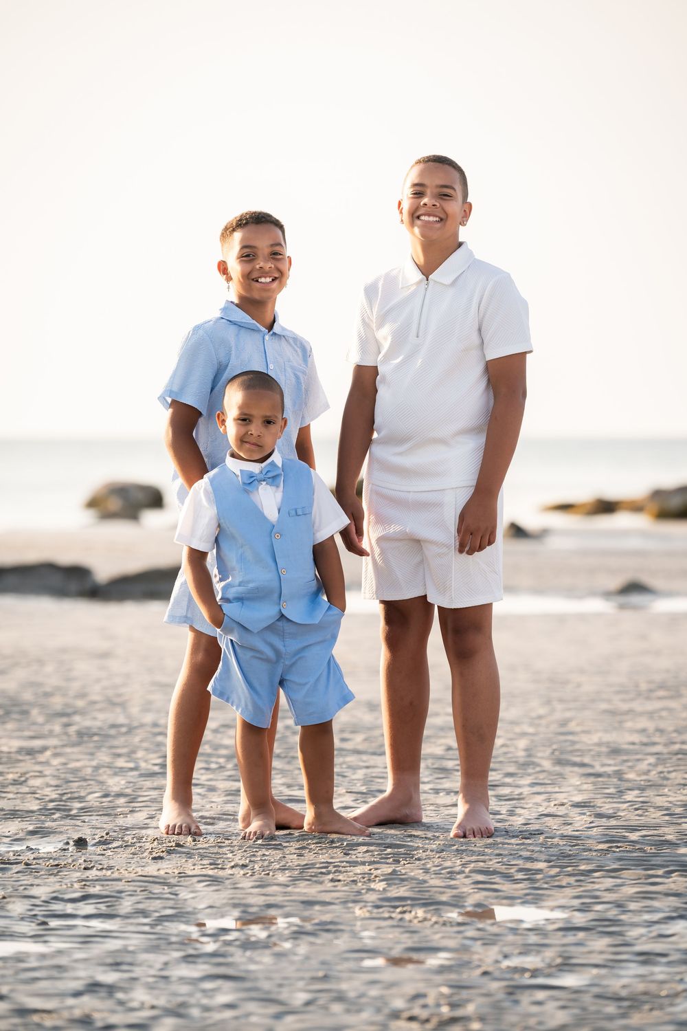 Three young boys in light blue and white outfits smiling together on a sandy beach at sunset.