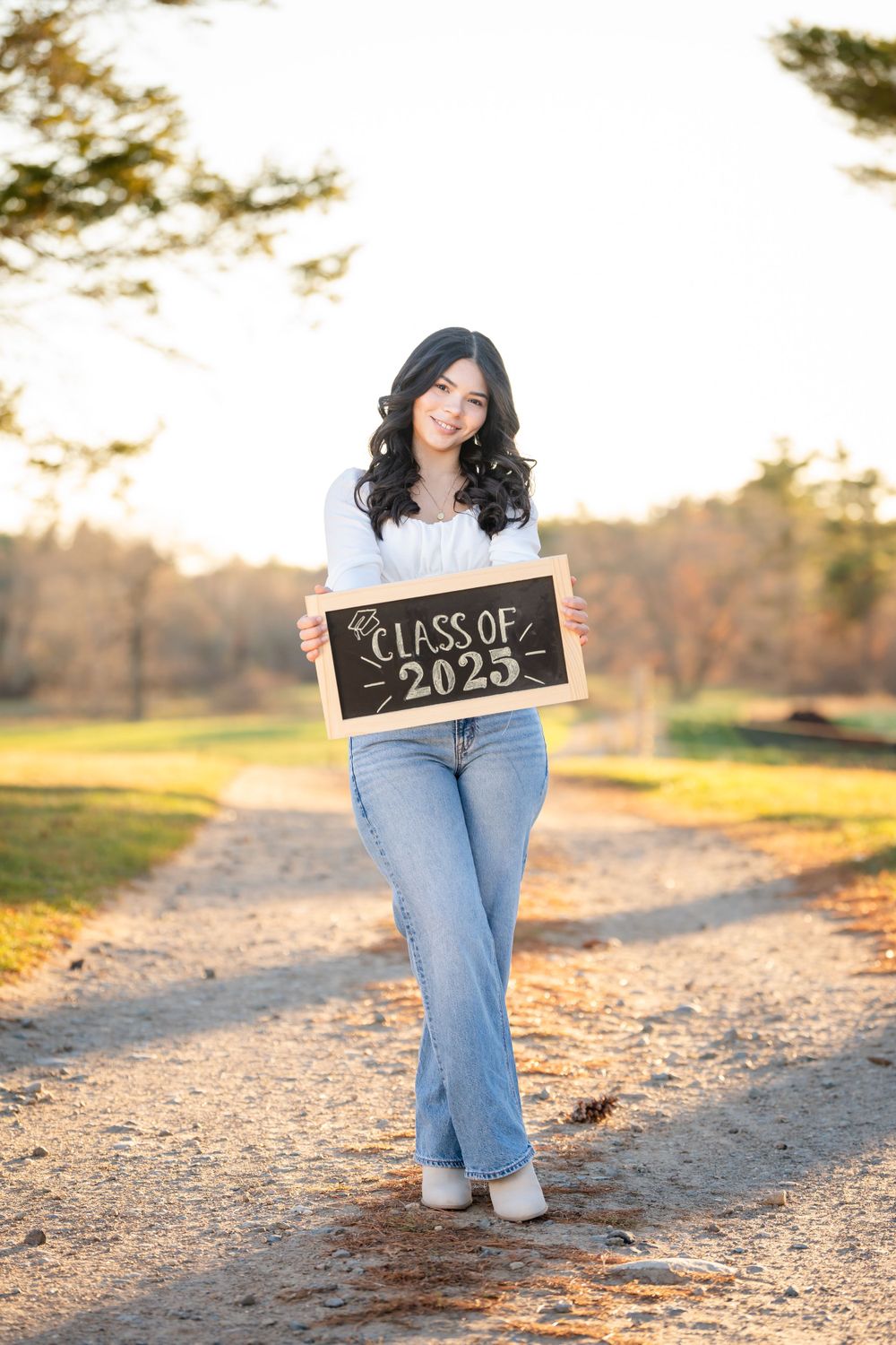 A graduate holds a 'Class of 2023' chalkboard sign while standing on a dirt path during golden hour sunset.