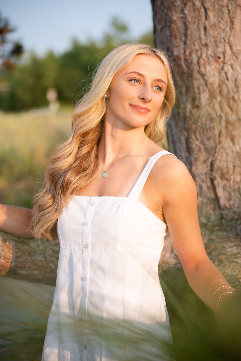 Young blonde woman in white sundress smiling outdoors near tree in golden sunlight.