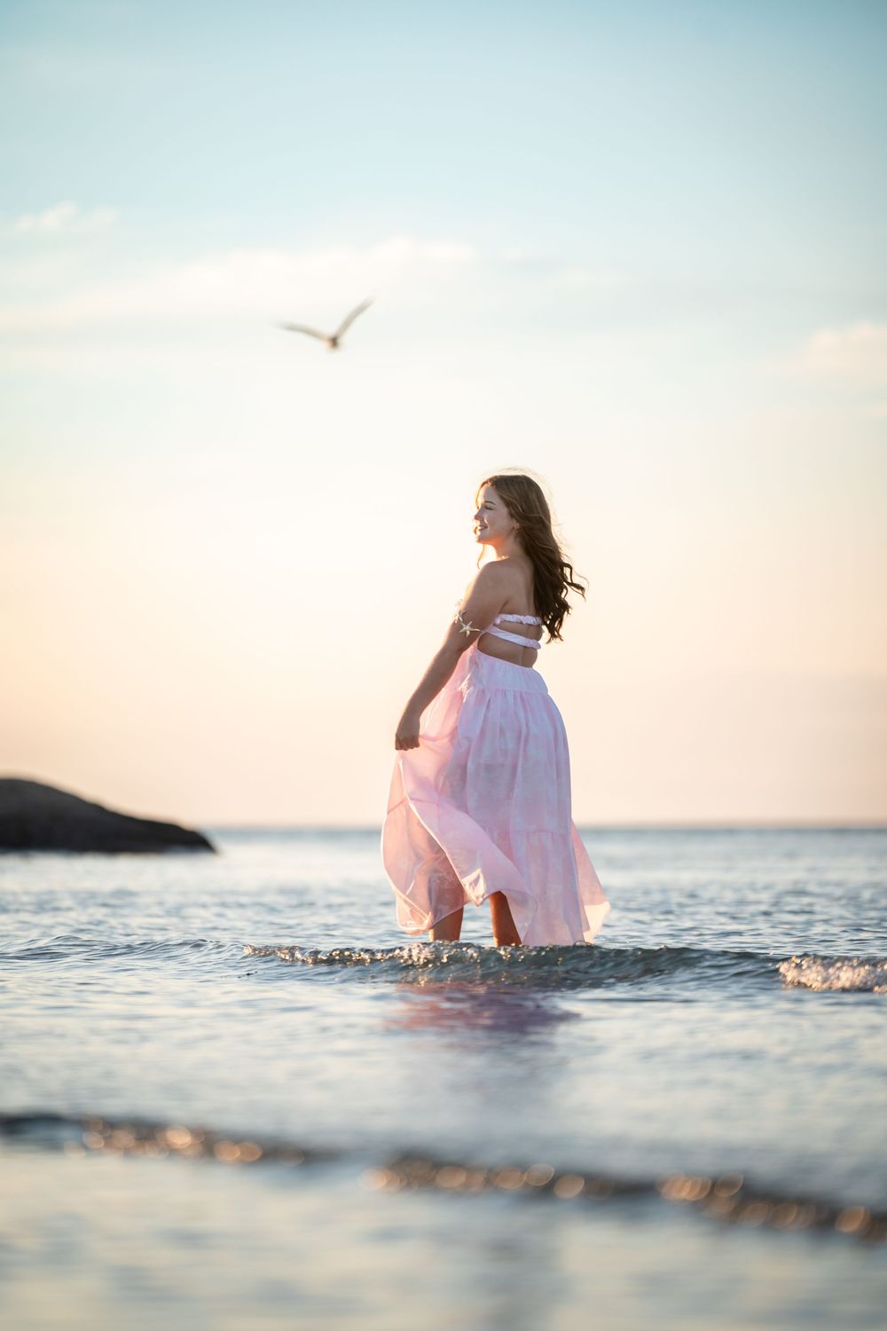 Woman in pink dress wading in ocean at sunset with seagull flying overhead.