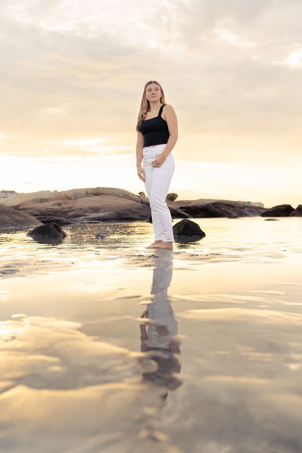 A person stands on wet beach rocks at sunset, creating a beautiful reflection in the shallow water.