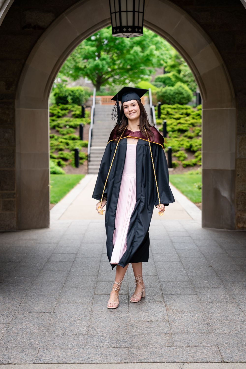A graduate stands in traditional cap and gown under a stone archway on a college campus with green trees in background.