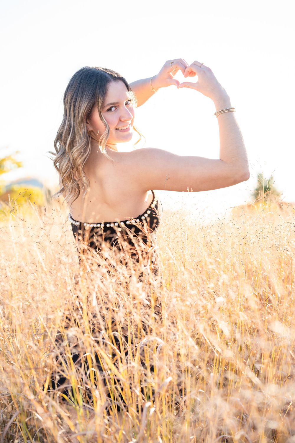 A woman in a leopard print dress makes a heart shape with her hands against golden sunlit grass in a field.