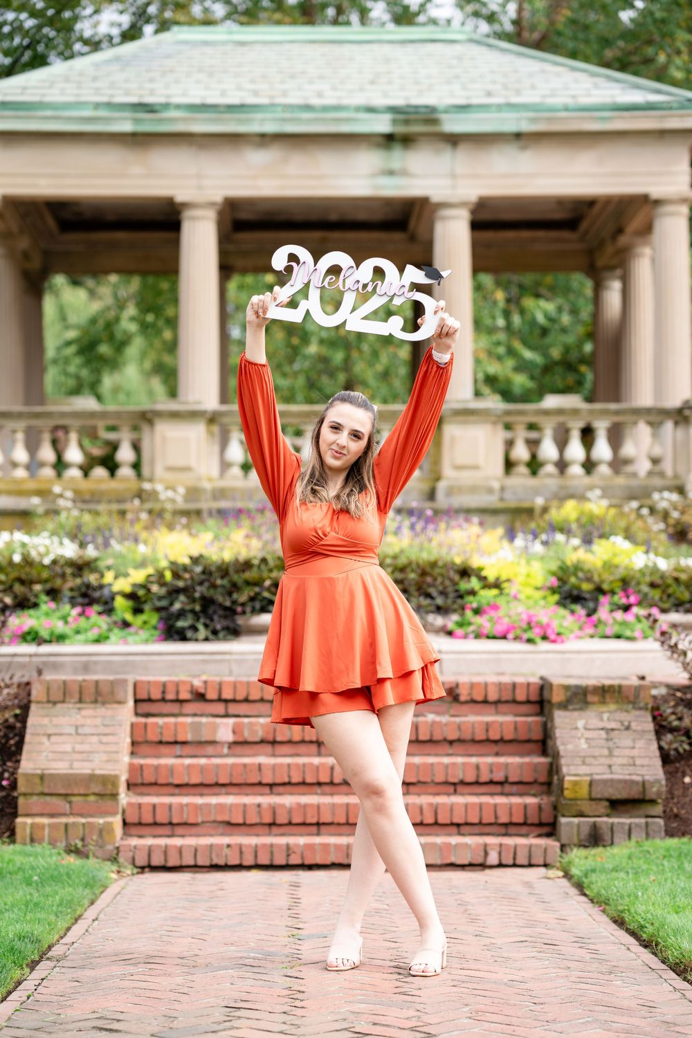 Graduate celebrates in a vibrant orange dress at a garden gazebo with decorative 2023 sign.