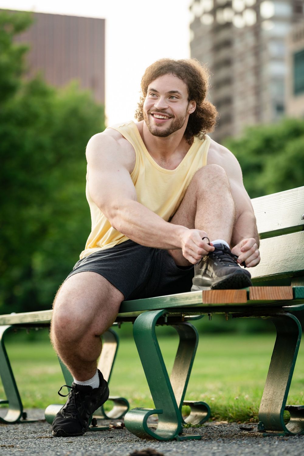 Muscular man in yellow tank top tying sneakers on park bench, smiling outdoors with city buildings in background.