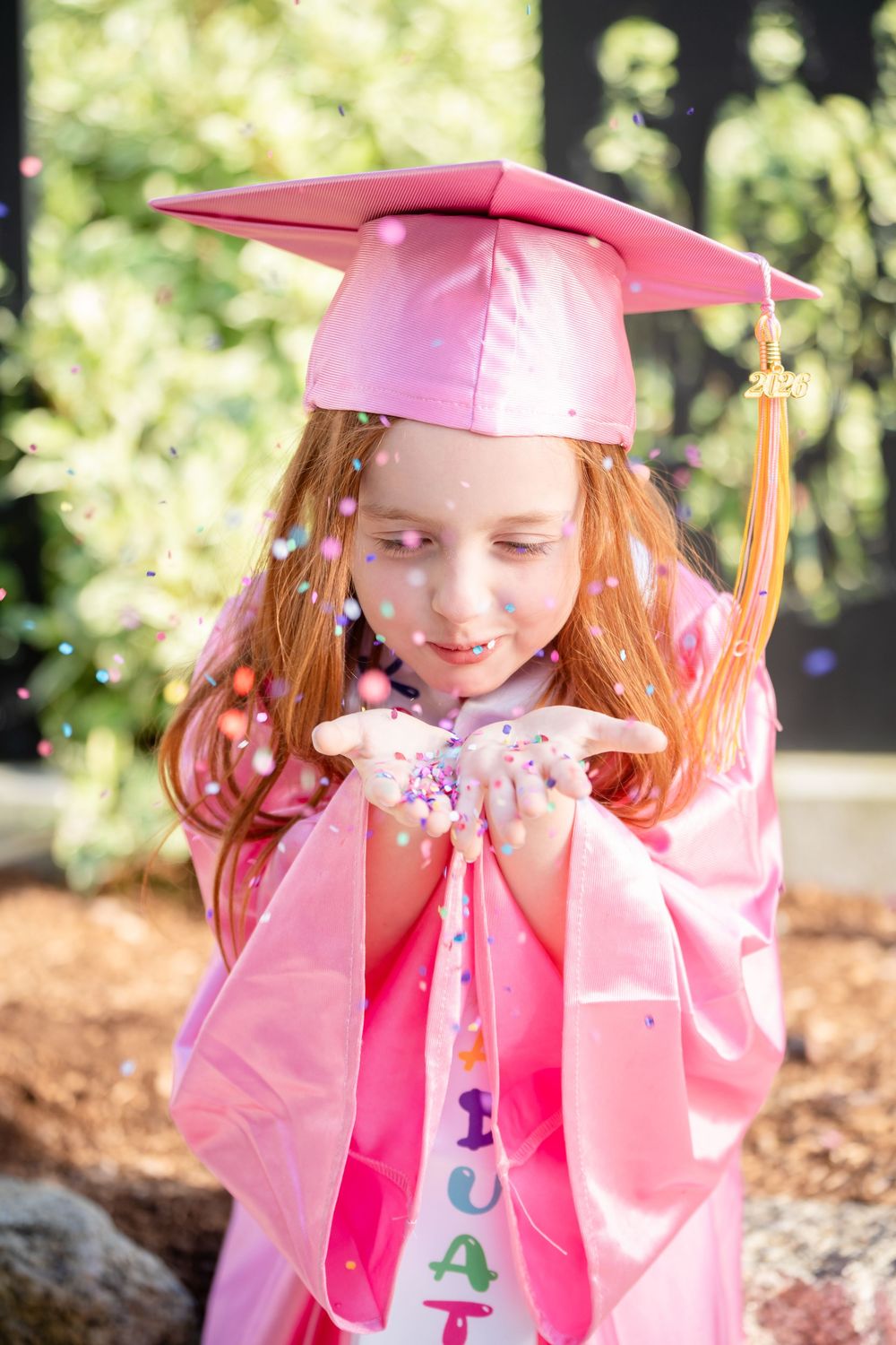 Young girl in pink graduation cap and gown blowing colorful confetti from her hands outdoors.