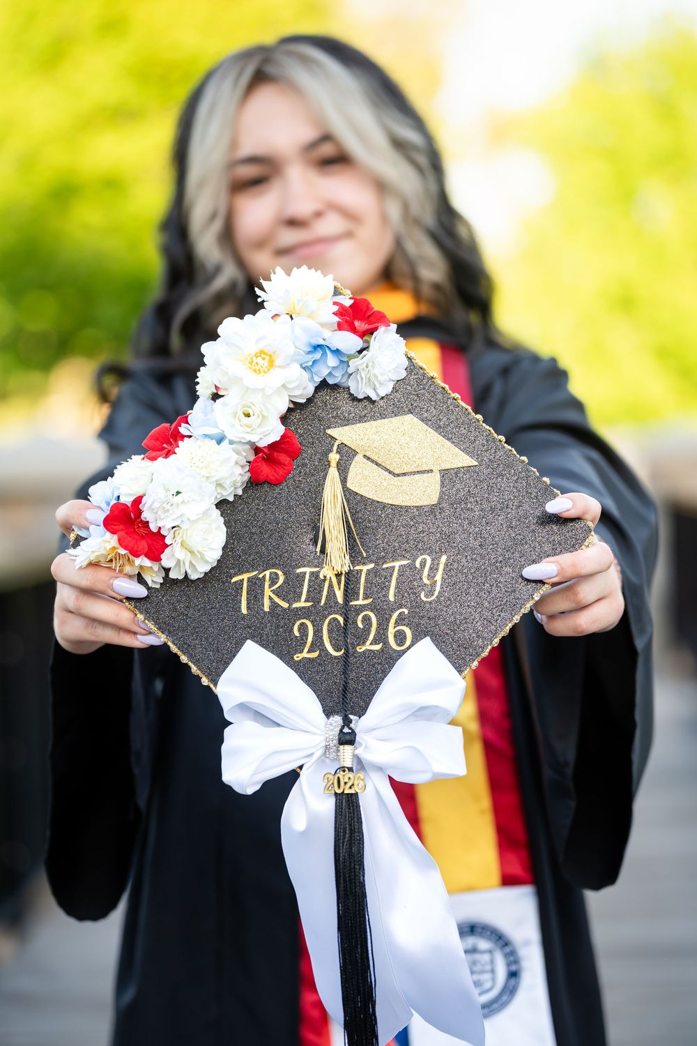 A graduate proudly displays a decorated mortarboard cap reading Trinity 2026 adorned with colorful flowers and a white bow.