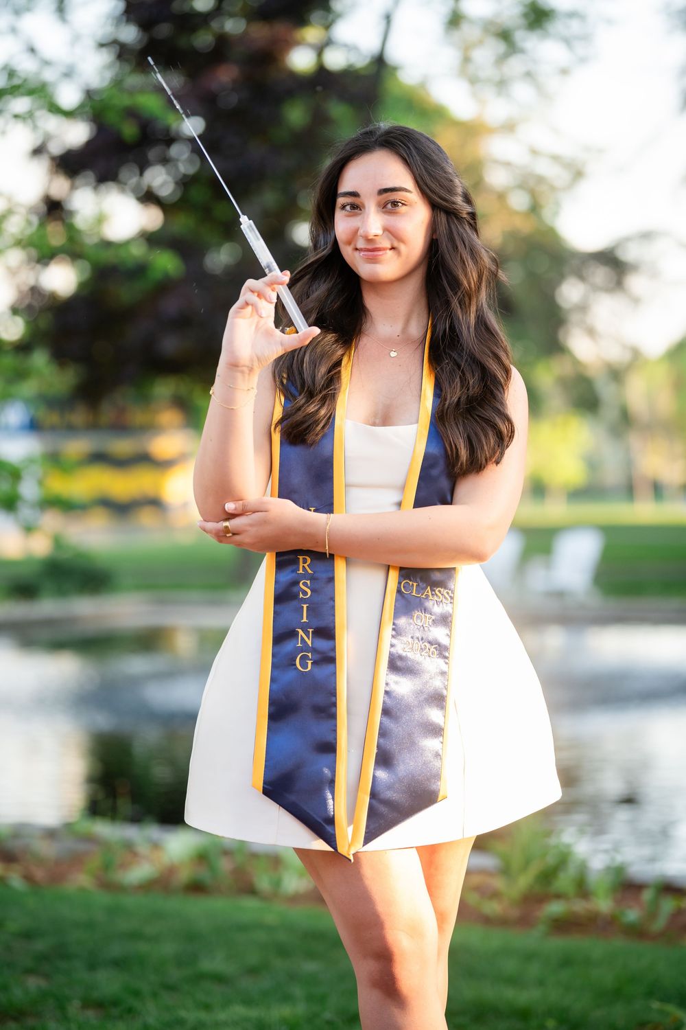 Nursing graduate smiles holding a syringe wearing a navy and gold graduation stole outdoors.