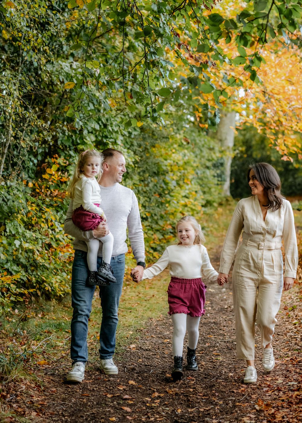A happy family of four enjoys a walk in a sunlit park, surrounded by lush greenery. Family photoshoot in Ireland.