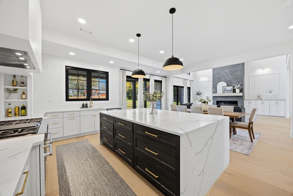 Modern kitchen with black island, gold hardware, marble countertops, white cabinets, pendant lights, and open living area.