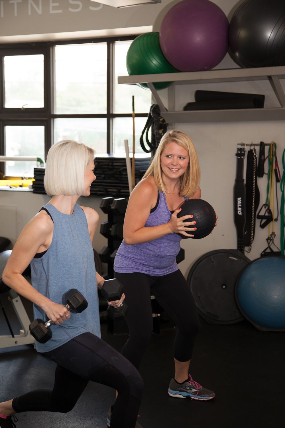 People working out with dumbbells and medicine balls in a fitness studio with colorful exercise balls on wall shelves.